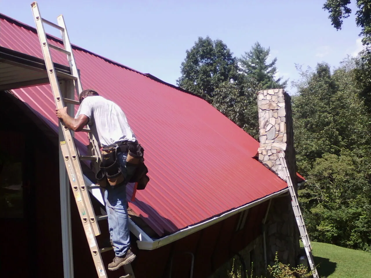 Roofer on a ladder beside a red metal roof and stone chimney — Soffit & Fascia Repair by Roofing Brothers in Lafayette Hill